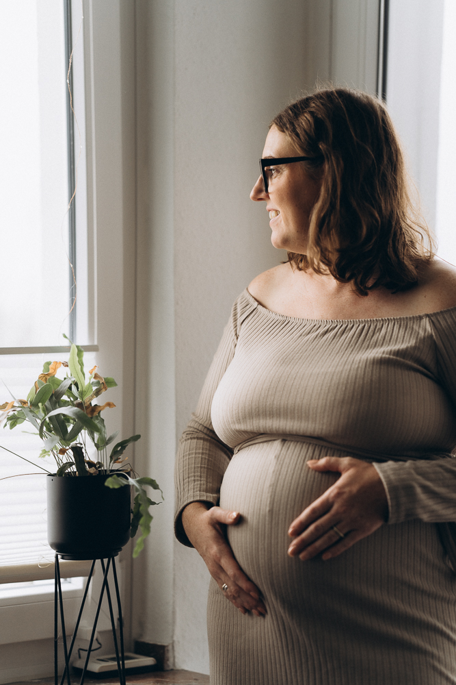 Babybauchshooting Mama schaut verträumt aus dem Fenster und streichelt den Bauch